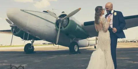 A bride and groom standing together in front of a vintage propeller airplane.