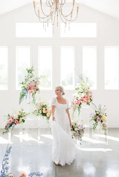A bride in a white lace dress stands in a bright, white-walled venue with large floral arrangements and a chandelier.