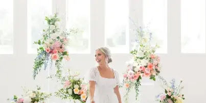 A bride in a white lace dress stands in a bright, white-walled venue with large floral arrangements and a chandelier.