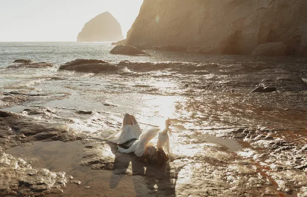 A person in a white dress lying on a rocky shoreline with the sun reflecting off the wet rocks and water.