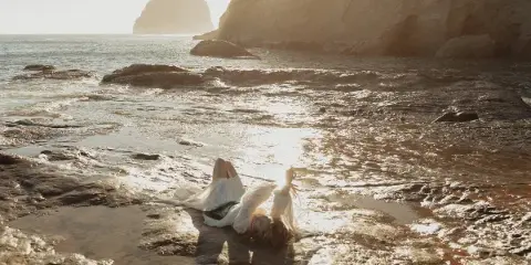 A person in a white dress lying on a rocky shoreline with the sun reflecting off the wet rocks and water.