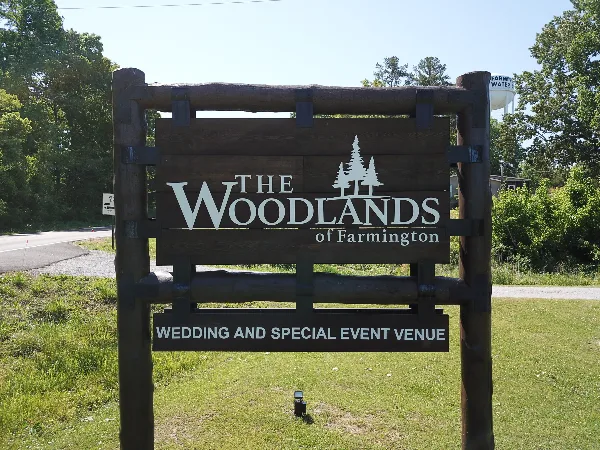 A wooden outdoor sign reading 'The Woodlands of Farmington Wedding and Special Event Venue' set against a grassy background with trees.
