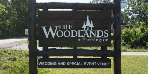 A wooden outdoor sign reading 'The Woodlands of Farmington Wedding and Special Event Venue' set against a grassy background with trees.