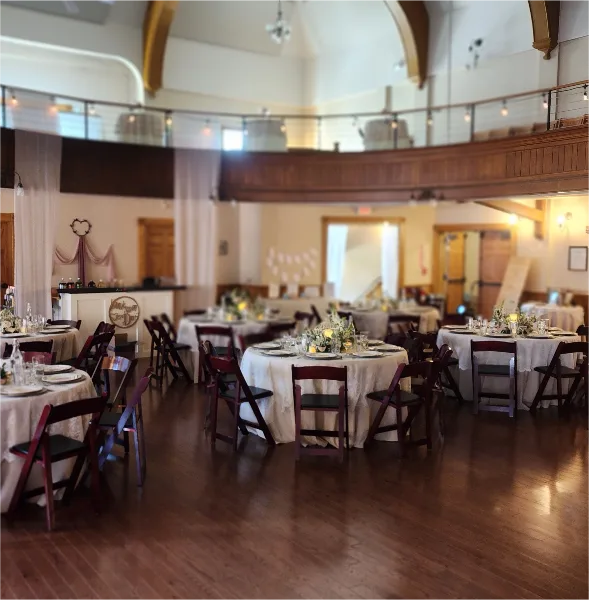 Interior view of a large event hall with a balcony, featuring round tables set with white linens and floral centerpieces for a wedding reception.
