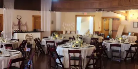 Interior view of a large event hall with a balcony, featuring round tables set with white linens and floral centerpieces for a wedding reception.
