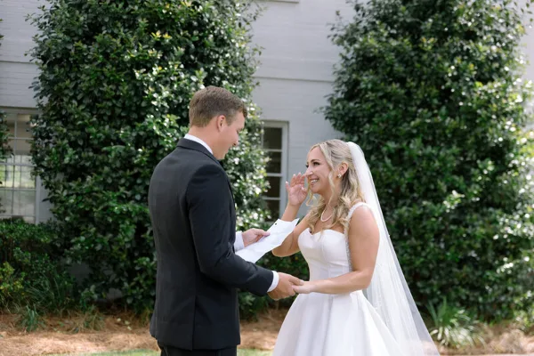 A bride and groom exchanging vows during an outdoor wedding ceremony in front of a white building with greenery.