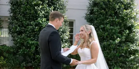 A bride and groom exchanging vows during an outdoor wedding ceremony in front of a white building with greenery.