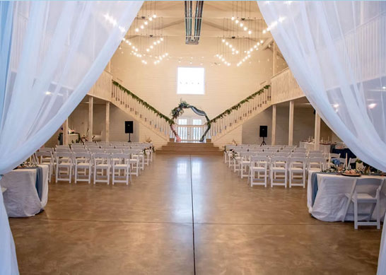 Interior view of a large white event barn set up for a wedding ceremony with rows of chairs and a floral arch.