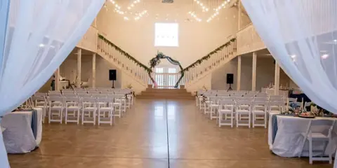 Interior view of a large white event barn set up for a wedding ceremony with rows of chairs and a floral arch.