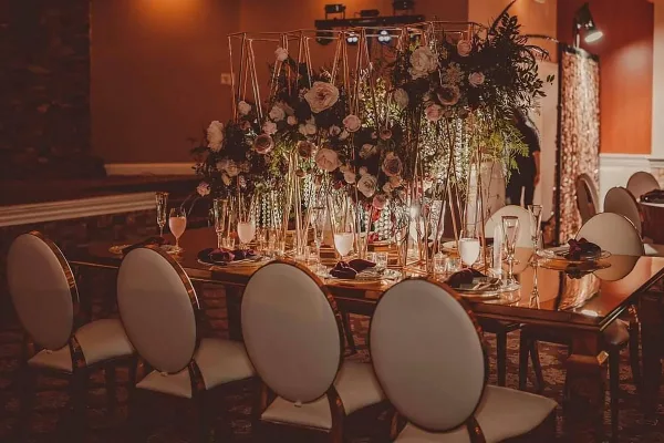 A luxury event table setting featuring upholstered chairs, glassware, and a large floral centerpiece in a geometric frame.