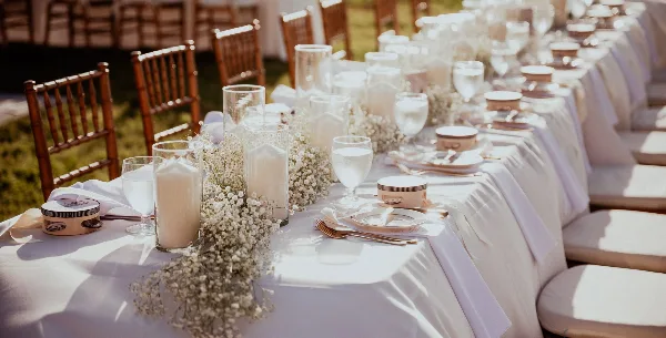 A long outdoor event table set with white linens, glassware, candles, and baby's breath floral arrangements.