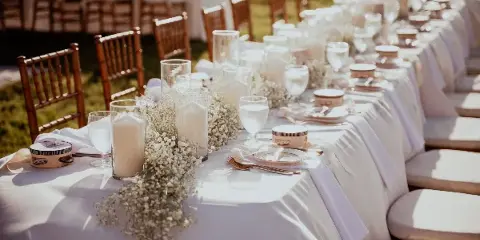 A long outdoor event table set with white linens, glassware, candles, and baby's breath floral arrangements.