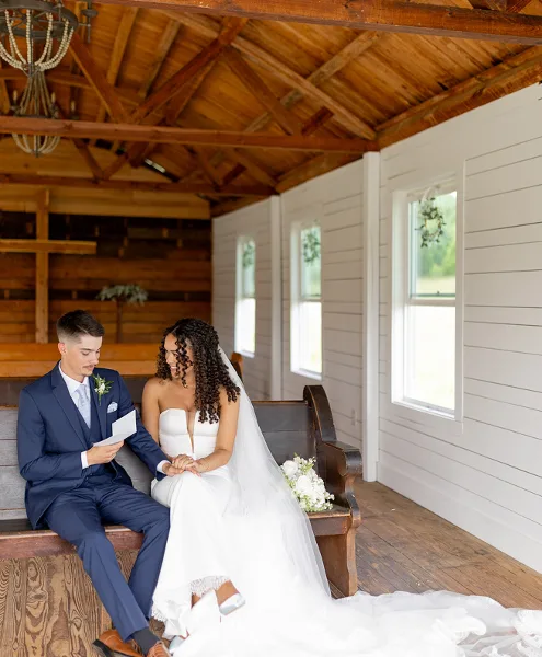 Bride and groom seated on a wooden bench inside a rustic chapel with exposed beams and a cross.