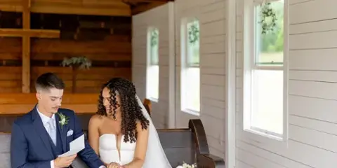 Bride and groom seated on a wooden bench inside a rustic chapel with exposed beams and a cross.