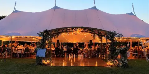 A large white wedding tent at dusk with string lights, floral decorations, and a dance floor.