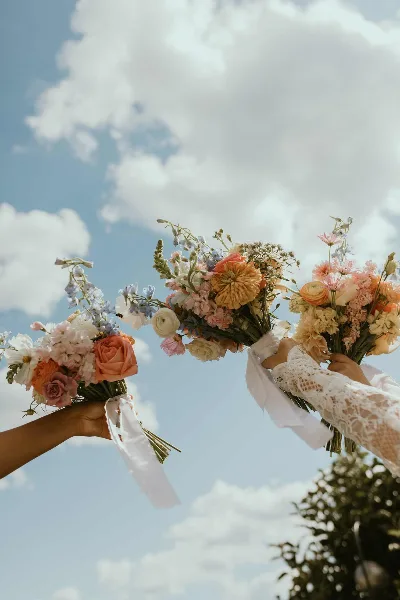 Three hands holding colorful, fresh flower bouquets against a blue sky with white clouds.