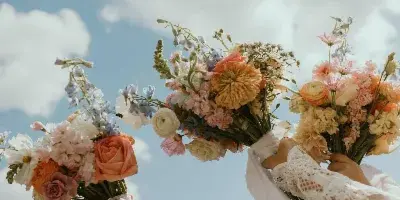 Three hands holding colorful, fresh flower bouquets against a blue sky with white clouds.
