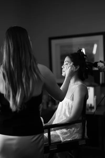 A black and white photograph of a makeup artist applying makeup to a bride seated in a director's chair.