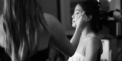 A black and white photograph of a makeup artist applying makeup to a bride seated in a director's chair.