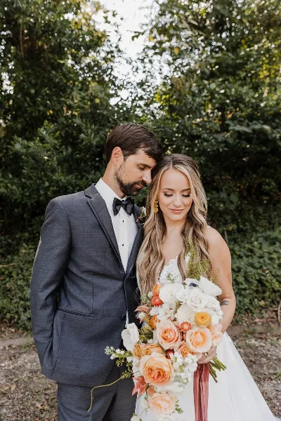 A bride and groom standing together outdoors, with the bride holding a large bouquet of peach, white, and orange roses and greenery.