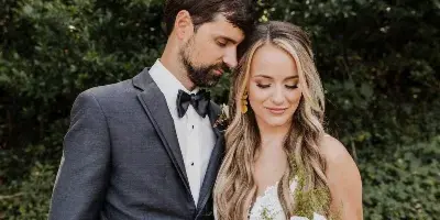 A bride and groom standing together outdoors, with the bride holding a large bouquet of peach, white, and orange roses and greenery.