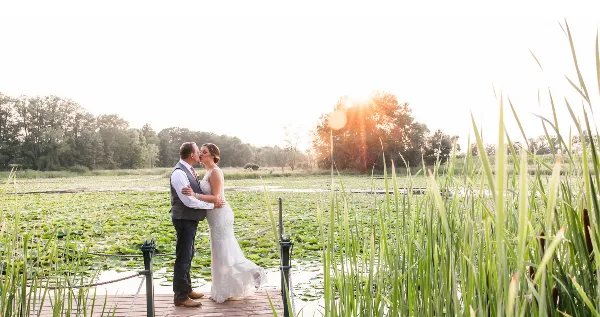 A bride and groom kissing on a wooden dock at a pond with tall reeds and a sunset in the background.