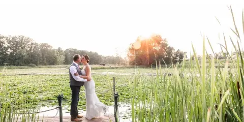 A bride and groom kissing on a wooden dock at a pond with tall reeds and a sunset in the background.