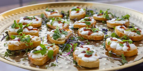 A gold serving tray filled with bruschetta topped with white cheese, microgreens, and a red sauce drizzle.