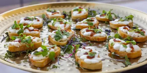 A gold serving tray filled with bruschetta topped with white cheese, microgreens, and a red sauce drizzle.