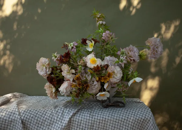 A floral arrangement with pink peonies and white ranunculus in a vase on a checkered cloth.