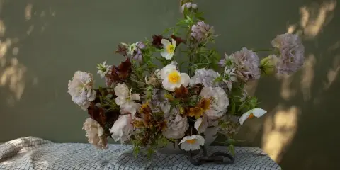 A floral arrangement with pink peonies and white ranunculus in a vase on a checkered cloth.