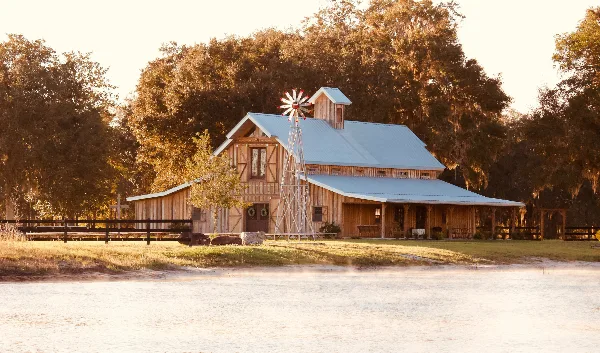 A rustic wooden barn with a metal roof and windmill situated next to a pond at sunset.
