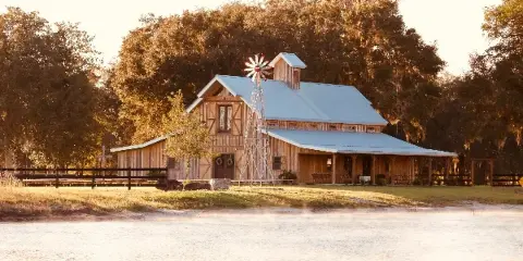 A rustic wooden barn with a metal roof and windmill situated next to a pond at sunset.