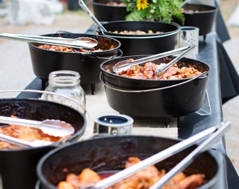 A table spread featuring several black cast-iron Dutch ovens filled with cooked food and serving spoons.