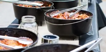 A table spread featuring several black cast-iron Dutch ovens filled with cooked food and serving spoons.