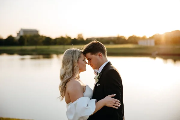 A bride and groom embracing in front of a lake at sunset.