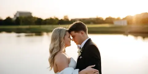 A bride and groom embracing in front of a lake at sunset.