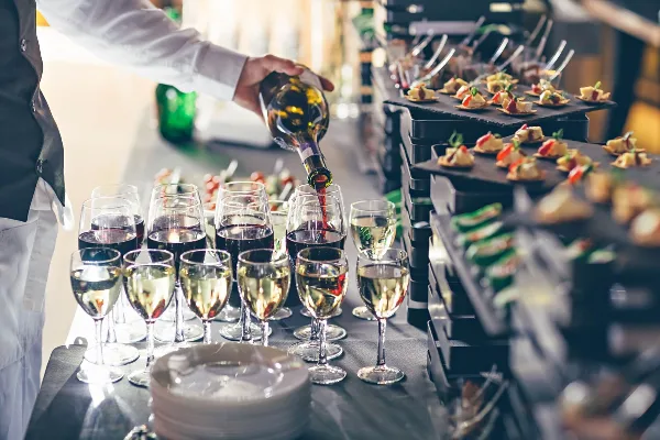 A server in a white shirt and black vest pours red wine into glasses next to a tiered display of gourmet appetizers.