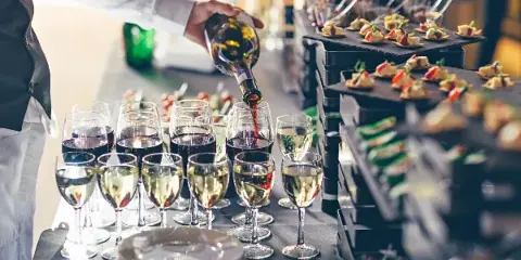 A server in a white shirt and black vest pours red wine into glasses next to a tiered display of gourmet appetizers.