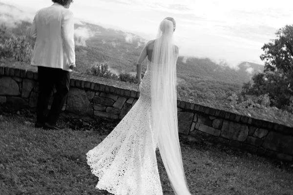 Black and white photograph of a bride in a lace gown and long veil standing with a groom in a white jacket, overlooking a misty mountain valley.