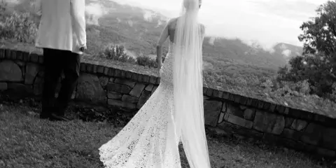 Black and white photograph of a bride in a lace gown and long veil standing with a groom in a white jacket, overlooking a misty mountain valley.