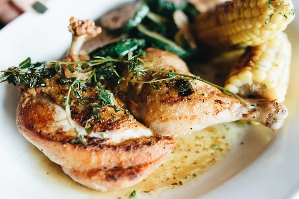 A close-up photograph of a plated meal featuring roasted chicken legs garnished with fresh herbs, served alongside roasted corn and vegetables.