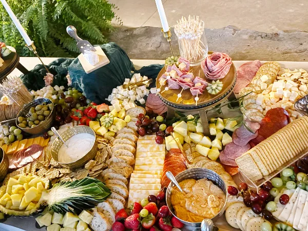 A large catering spread featuring a charcuterie board with sliced meats, cheeses, grapes, strawberries, pineapple, bread, crackers, and dips arranged on a table.
