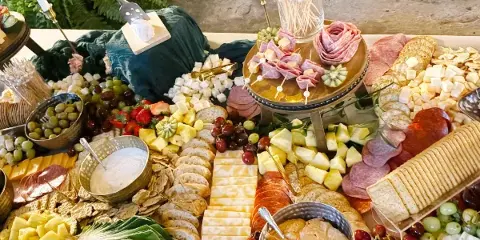 A large catering spread featuring a charcuterie board with sliced meats, cheeses, grapes, strawberries, pineapple, bread, crackers, and dips arranged on a table.