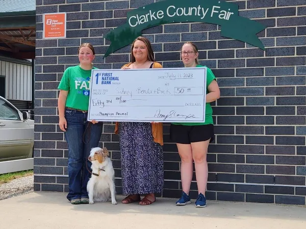 Three people holding a large check in front of a brick wall with a 'Clarke County Fair' sign and a dog sitting at their feet.
