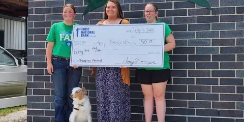 Three people holding a large check in front of a brick wall with a 'Clarke County Fair' sign and a dog sitting at their feet.