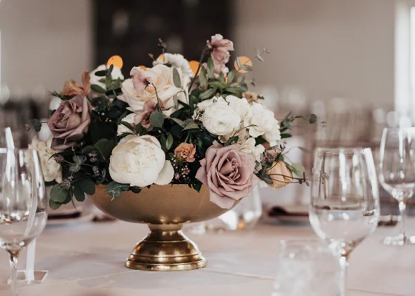 Wedding table centerpiece with white and blush roses in a gold bowl, surrounded by wine glasses on a set table.
