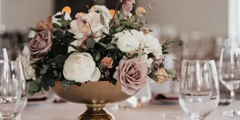 Wedding table centerpiece with white and blush roses in a gold bowl, surrounded by wine glasses on a set table.