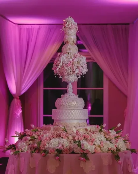 A tall, ornate white wedding cake with elaborate sugar flower decorations and a floral base, displayed on a table under pink lighting.