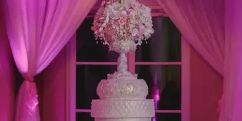 A tall, ornate white wedding cake with elaborate sugar flower decorations and a floral base, displayed on a table under pink lighting.